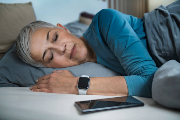 aplicativos de sono - mulher sênior experiente em tecnologia dormindo na cama, usando telefone inteligente e relógio inteligente para melhorar seus hábitos de sono - tracker - fotografias e filmes do acervo