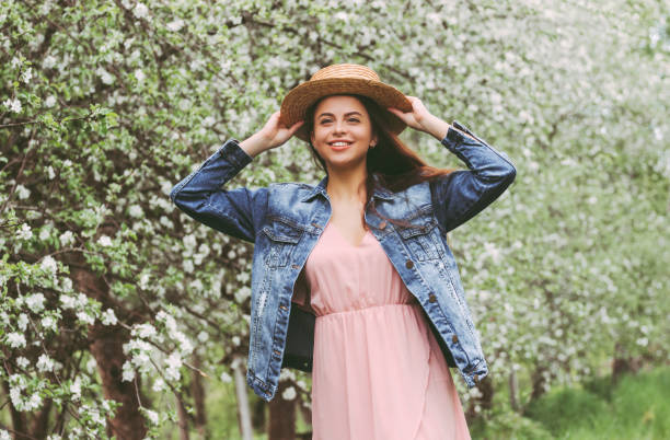 Cheerful young woman in straw hat and dress have fun outdoors Portrait happy carefree brunette woman hipster have fun on green garden with blossom flower trees. Young beautiful hippie girl in stylish straw hat and pink dress relaxing on spring countryside meadow denim jacket stock pictures, royalty-free photos & images