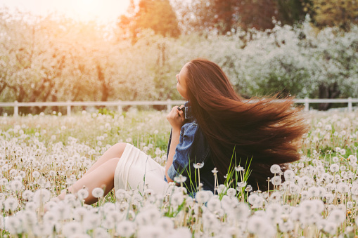 Back view beautiful carefree hipster woman posing with her long healthy hair in motion on blossom countryside floral garden. Young attractive hippie girl in denim jacket sit on dandelion flower field