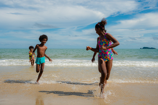 Group of African American boys and girls in swimsuit run from the sea to beach under beautiful blue sky. Concept of outdoor activity in summer after covid 19 lock down measure