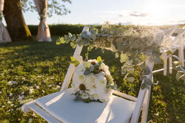 Bright white bridal bouquet on a chair standing on a meadow, in the evening backlight with sunset