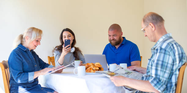 朝食を食べながら午前中にニュースを読む幸せな多世代家族は、若者に焦点を当てる - デジタル認知症とデジタル技術コンセプトの過剰使用 - young adult reading newspaper the media ストックフォトと画像