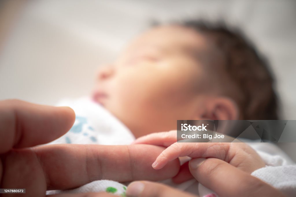 A newborn infant baby girl in a blanket swaddle wraps her tiny hand and fingers around her father and mother's fingers as she sleeps peacefully. Newborn Stock Photo