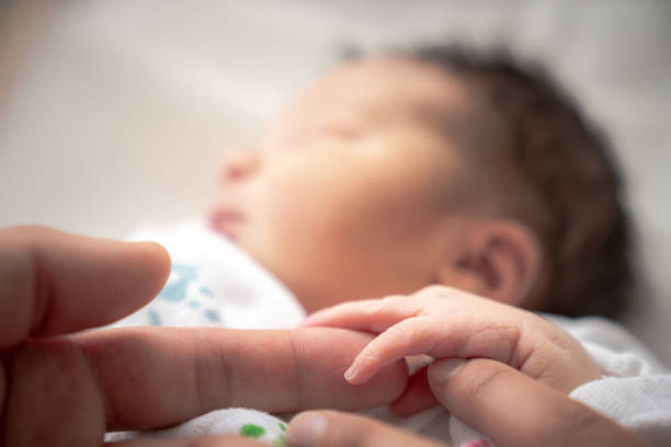 a newborn infant baby girl in a blanket swaddle wraps her tiny hand and fingers around her father and mother's fingers as she sleeps peacefully. - baby mother sleeping child imagens e fotografias de stock