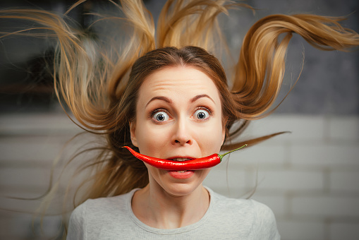 A girl with a red hot pepper in her teeth. Chili for cooking. The spicy seasoning is very stinging in the mouth. My hair stood on end. Bite the pepper and get burned. Emotional woman.