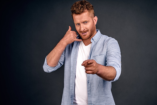 Young attractive guy with red hair in casual shirt, makes phone sign, indicates with index finger directly at camera, wants to speak with someone via cell phone. Body language concept.