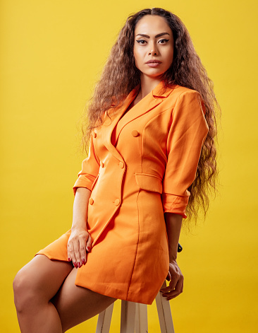 Beautiful woman with long and curly hair in orange jacket sitting on chair front of yellow background.