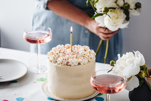 Birthday cake with wine glasses on table. Birthday Cake with a lit candle and a woman in background holding flowers.