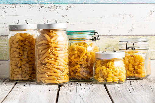 Various types of raw italian pasta in jars on wooden table.