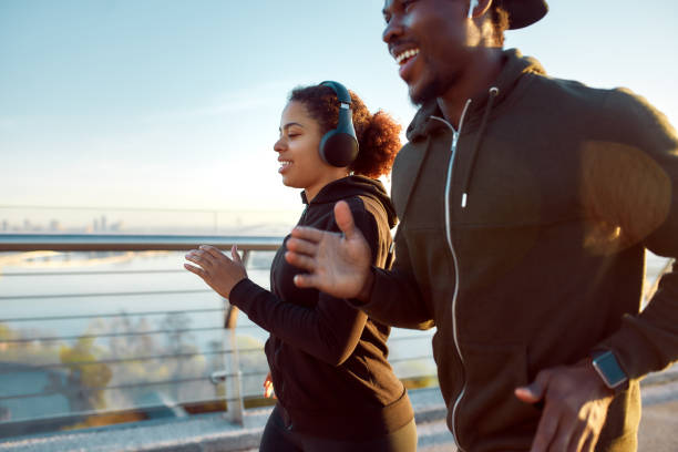 朝のスポーツ。橋の上で一緒にジョギングヘッドフォンで若い笑顔のアフリカのカップル。屋外で走って楽しんで幸せなアフリカの男女 - running jogging african descent nature ストックフォトと画像
