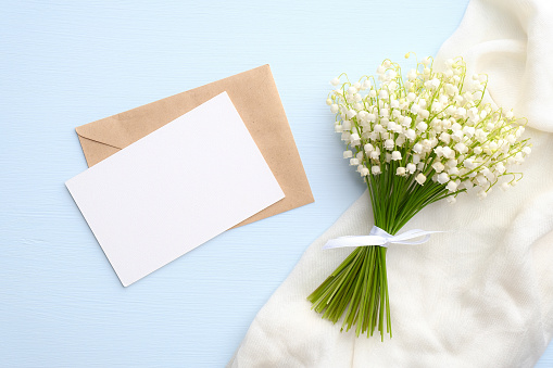 Bouquet of flowers lily of the valley and blank paper card with envelope on blue table from above. Beautiful vintage postcard, top view, space for text, flat lay.