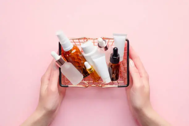 Female hands holding shopping basket with cosmetics on pink background. Beauty products sale concept.