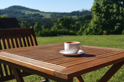 Cup of coffe on wooden table with chair. Blurred green trees and hill tops background. Coffee break in countryside. Picnic, garden party snack, relaxation concept,no people, selective focus.
