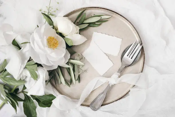 Photo of Festive summer wedding scene. Marble table setting with cutlery, olive branches, white peony flowers, stoneware plate and silk ribbon. Blank place cards mockups. Restaurant menu concept. Flat lay, top