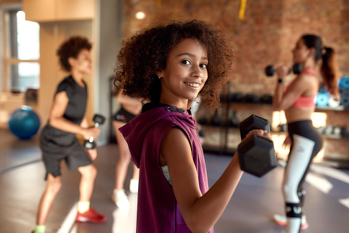 African american girl smiling at camera while exercising using dumbbell in gym together with female trainer and other kids. Sport, physical education concept. Horizontal shot. Selective focus