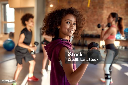 istock African american girl smiling at camera while exercising using dumbbell in gym together with female trainer and other kids. Sport, healthy lifestyle, physical education concept 1247981111