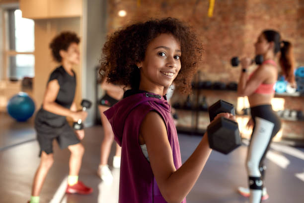 African american girl smiling at camera while exercising using dumbbell in gym together with female trainer and other kids. Sport, healthy lifestyle, physical education concept African american girl smiling at camera while exercising using dumbbell in gym together with female trainer and other kids. Sport, physical education concept. Horizontal shot. Selective focus aerobics photos stock pictures, royalty-free photos & images