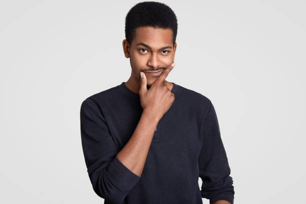 istock Hesitant black man holds chin, looks doubtfully, purses lips in uncertainty, has short curly hair, wears casual clothes, tries make decision, isolated over white wall. Contemplative dark skinned guy 1248064884