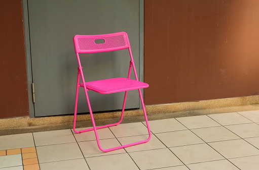 Closeup empty hot pink folding chair placed in front of closed gray door and brown wall under sunlight