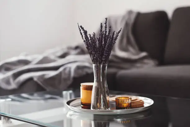 Photo of Living room decor: glass coffee table with dried lavender bouquet and aroma candles on a metal tray, sofa and blanket on background. Scandinavian style. Simple home decoration