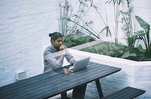Capturing the individuality of a young bearded man, this portrait showcases him working on a laptop outdoors in the city. His handsome and stylish appearance,