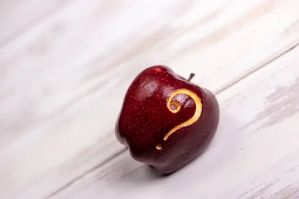 Red shiny delicious apple with an question mark sign carved into it, photographed from the side while rolled/placed on white painted aged pine timber floor boards with pin sharp focus on the fruit. Ideal for many concept with plenty of copy space for your message and ideas, but mainly aiming to represent healthy eating, an "alertness" for nutrition - importance of nutrition, ripe fruit, healthy lifestyle, question sigh, healthcare and medicine, carving - craft product, etc. Shot on Canon EOS R system in studio for highest quality and resolution.
