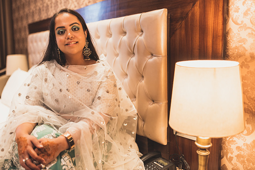Delhi, India - 01/01/2019: A rich Indian girl from a royal family sitting inside an sitting in a luxurious room next to warm lamp. Girl wearing traditional Indian clothing as she gets ready for a wedding ceremony.