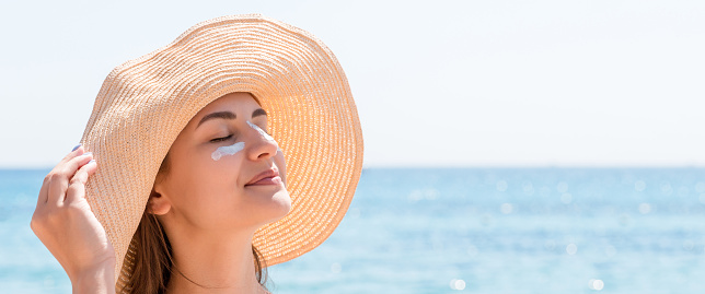 Beautiful young woman in hat is applying sunblock under her eyes and on her nose like Indian. Sun protection concept.