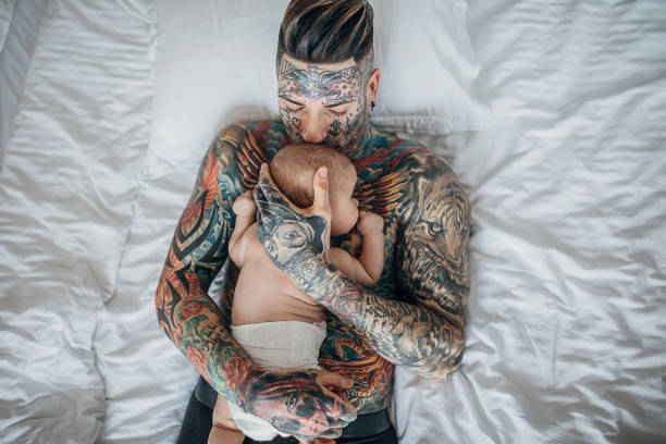 father with whole body covered in tattoos lying with his baby boy on bed in bedroom - tattoo father family son imagens e fotografias de stock