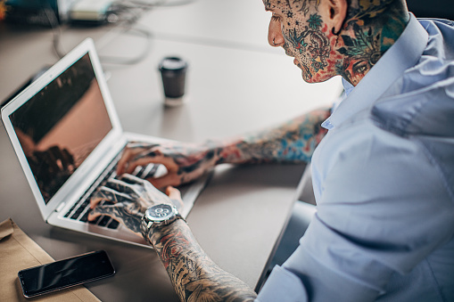 One young modern businessman with whole body covered in tattoos working in office, using laptop.