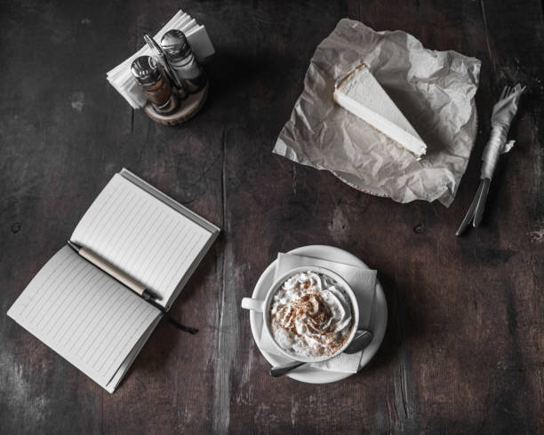 Dark photo. Cappuccino with whipped cream next to a notebook and cheesecake. Cozy coffee shop, selective focus