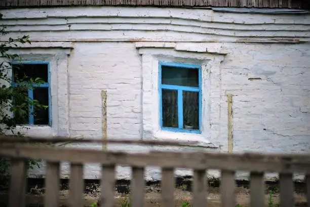 Photo of Facade of an old rural house with blue window frames. Old deformation of the wall structure caused by subsidence.