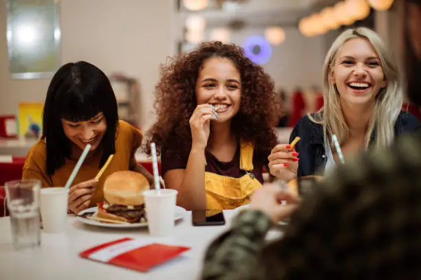 Three beautiful female friends laughing while eating burgers and fries in a restaurant