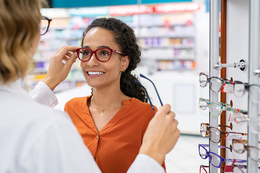African young woman in optic store choosing new cool-down glasses with optician. Mixed race girl trying new eyeglasses with the help of the pharmacist. Happy multiethnic woman trying new rest spectacle frame in optic store: eyesight and eye care concept.