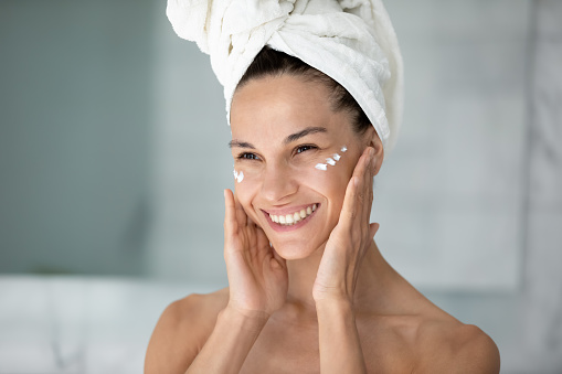 Close up head shot smiling young woman wearing white bath towel on head applying moisturizing face cream under eyes after shower, beautiful girl doing facial massage, enjoying perfect smooth skin