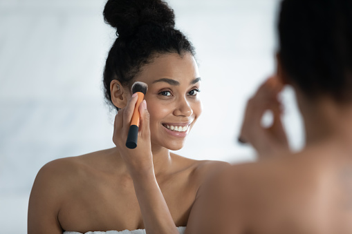 Head shot close up smiling African American young woman using cosmetics brush, looking in mirror, beautiful girl putting daily makeup, applying powder, standing in bathroom after shower