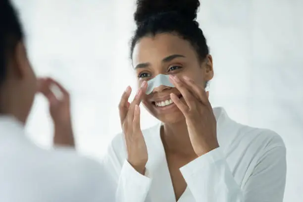 Mirror reflection close up smiling African American young woman applying anti blackhead patch, attractive girl wearing white bathrobe using nose strip mask, standing in bathroom, skincare procedure