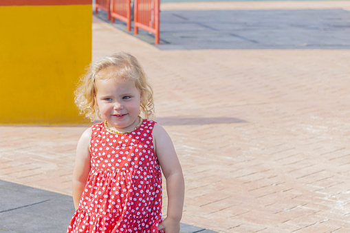 Cute little girl in red dotted dress walking on playground. One little girl walks on the playground in the street. Copy space. Summer recreation and happy childhood concept