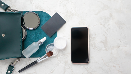 Concept of online purchase from smartphone in beauty fashion shop. Compact pocket mirror, facial serum, transparent powder, makeup brush and black credit card in bag on marble background, above