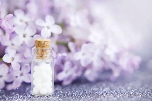 Homeopathic globules in glass bottle, close-up on a background of lilac flowers. Selective focus. Alternative medicine concept. Copy space