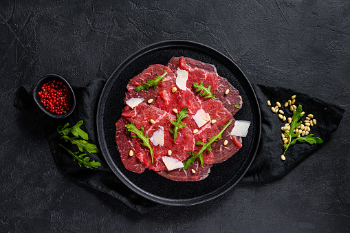 Marbled beef carpaccio with arugula and parmesan cheese. Black background. Top view. Space for text.
