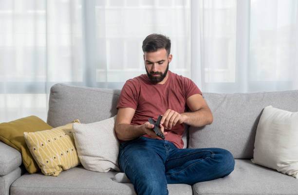 giovane bel hipster barbuto che apre il telecomando della tv per cambiare le batterie vuote o morte in modo che possa guardare il televisore dal divano nella sua stanza - watch battery foto e immagini stock