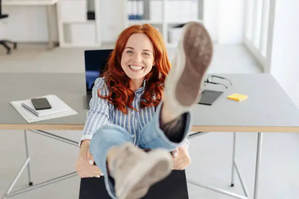 Carefree young businesswoman or student sitting with her legs over the back of her chair in the office grinning as she kicks her feet
