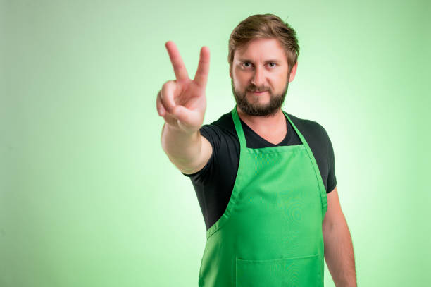 Supermarket employee with green apron showing victory Supermarket employee with green apron and black t-shirt, showing victory isolated on green background black men with blonde hair stock pictures, royalty-free photos & images