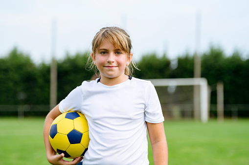 Close-up of 9 year old female footballer standing on field with ball under her arm and goal in background.