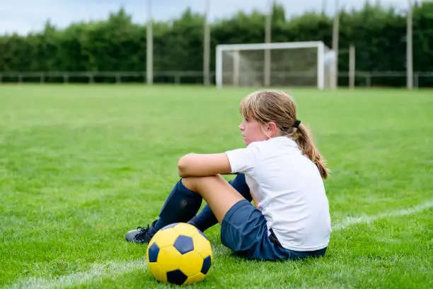 Close-up of serious young female footballer sitting on field sideline with ball and goal in background.