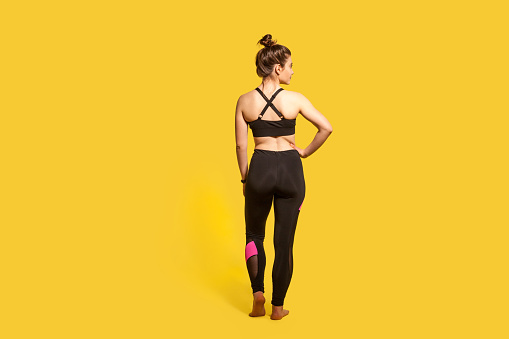 Fitness and sport training, daily workouts. Back view, slim sportswoman with hair bun in tight yoga pants showing her athletic body, trained back muscles. studio shot isolated on yellow background