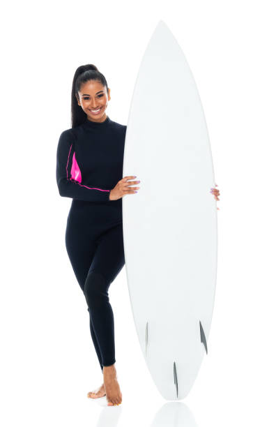 Front view of aged 20-29 years old who is beautiful with ponytail generation z female standing in front of black background wearing wetsuit who is happy who is barefoot and using surfboard