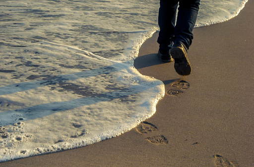 Foam from a sea wave reaching the shore or beach. Woman with boots walking on the sand, leaving footsteps next to the water. B