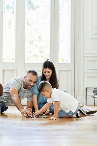 Woman looking at man and son playing with toy cars on floor. Happy family is enjoying quarantine together at home. They are sitting in living room.